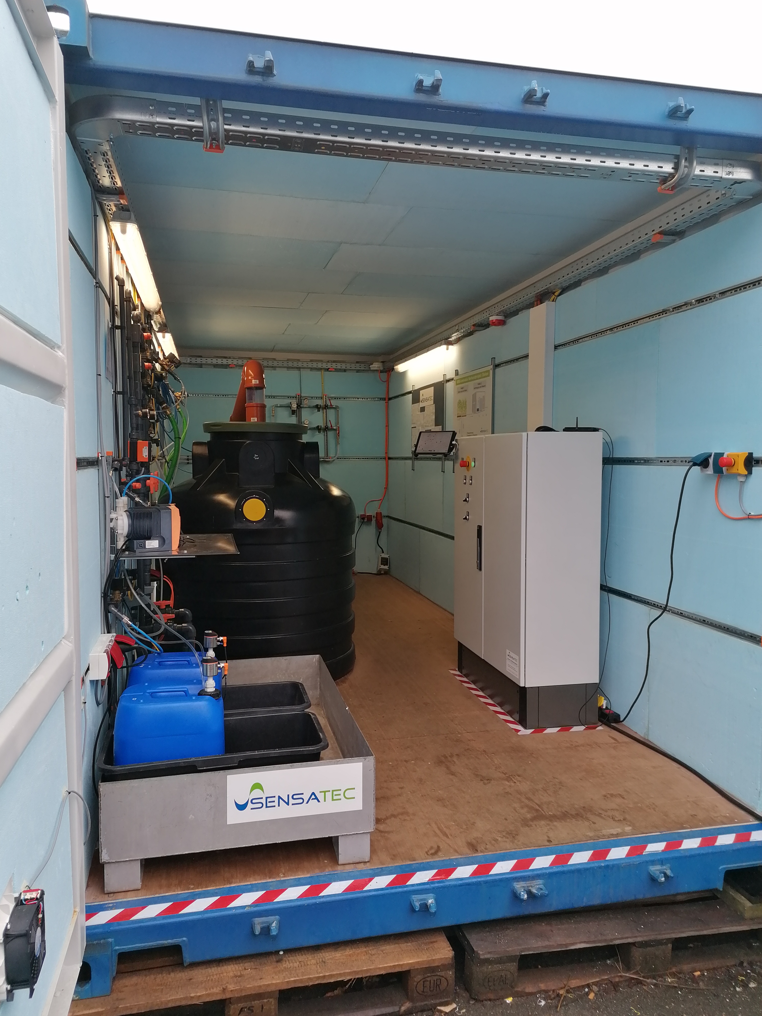 Das Bild zeigt die Innenansicht eines Seecontainers in welchem ein Schaltschrank steht, eine Dosier- und Verteilerstation bestehend aus Rohrleitungen und Pumpen sowie zwei große, schwarze Tanks (Flotationsbehälter) zu sehen sind.