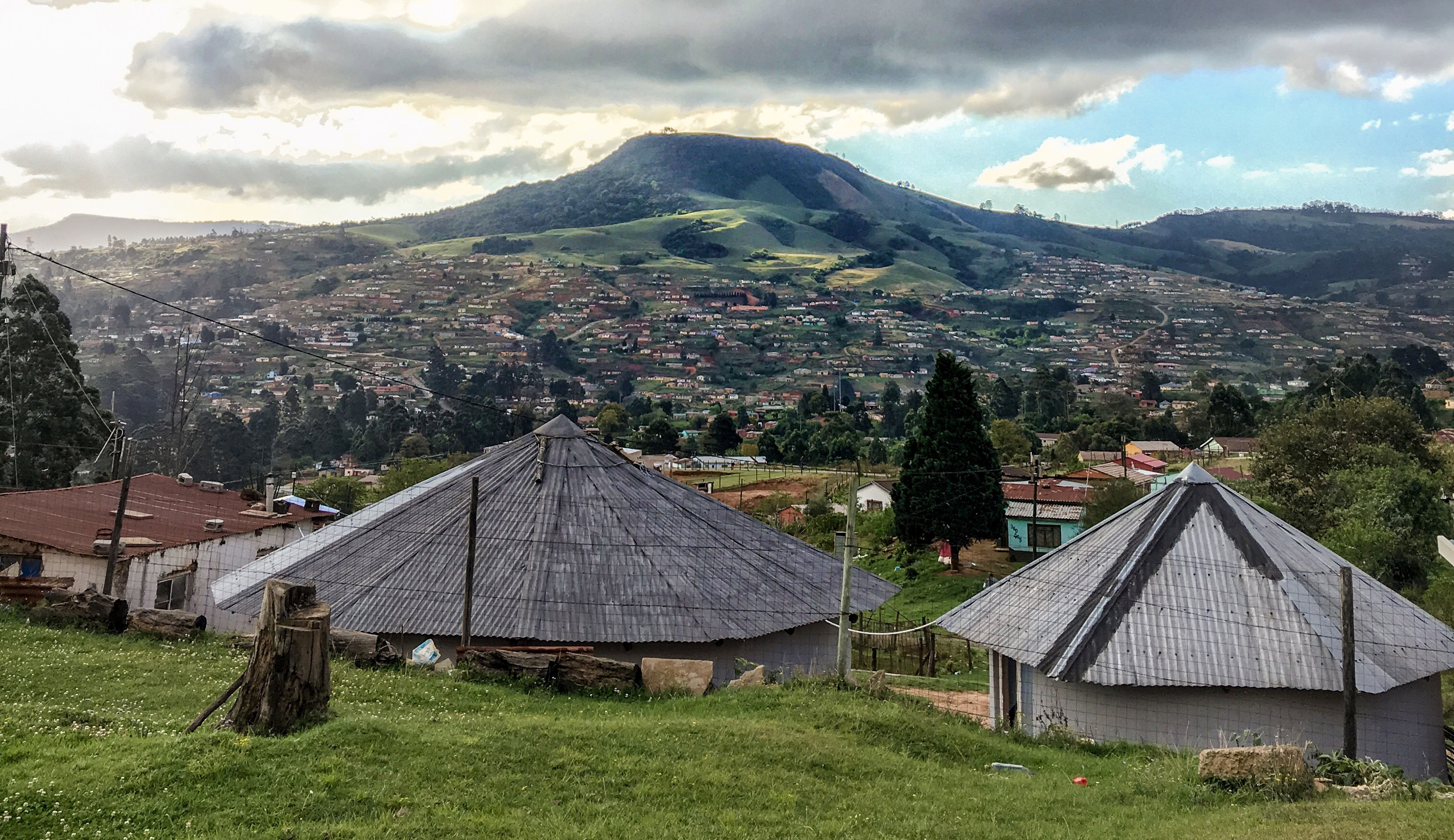 The picture shows a hilly landscape. A settlement partially covers the slopes. The hilltops are partly covered with forest. In the foreground, two traditional Zulu huts (round huts with pointed roofs) and a residential hut are visible.
