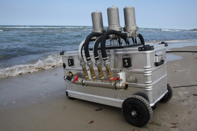 Side view of the 'Rocket'. An aluminum box contains a pump to draw water through filters. The four candle filters are each contained in a stainless steel enclosure and are mounted vertically on the box. They are connected to the pump and water inlet via hoses.