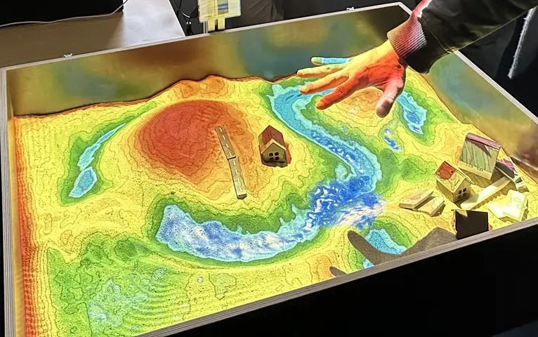 Im Sand sind Landschaftsformen wie Hügel und Täler modelliert, deren Höhenunterschiede durch eine farbige Projektion hervorgehoben werden. Das projizierte Geländemodell zeigt einen relativen Farbverlauf von Blau (unter dem Meeresspiegelniveau) über Grün, Gelb und Orange bis hin zu Rot und Braun für höher gelegene Bereiche. Die höchsten Darstellungsbereiche werden Weiß eingefärbt. Über dem Sand schwebt eine Hand, mit der Regen simuliert werden kann. Auf der Sandfläche ist zudem eine Wasseroberfläche sichtbar, die den simulierten Wasserfluss über das Gelände zeigt.
