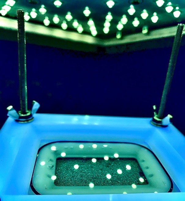 Reaktor im Labormaßstab in einer abgeschlossenen Bestrahlkammer, welche mit Schaumstoff verkleidet ist. An der Decke der Bestrahlkammer sind die LEDs angeordnet. Der Reaktor ist an vier Gewindestangen befestigt um den optimalen Abstand zu den LEDs gewährleisten zu können.